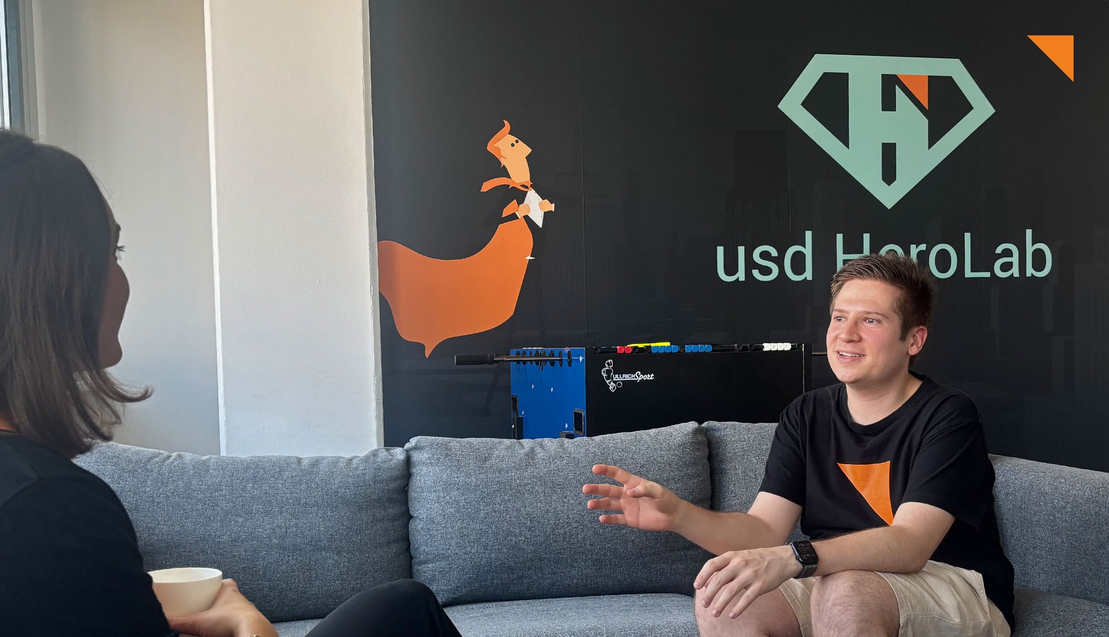 Mann sitzt auf einem Sofa und redet mit einer Frau. Im Hintergrund ist das Logo des usd HeroLabs zu sehen.